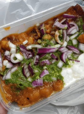 Chickpea curry topped with diced red onions, green chutney, and yogurt in a plastic container.