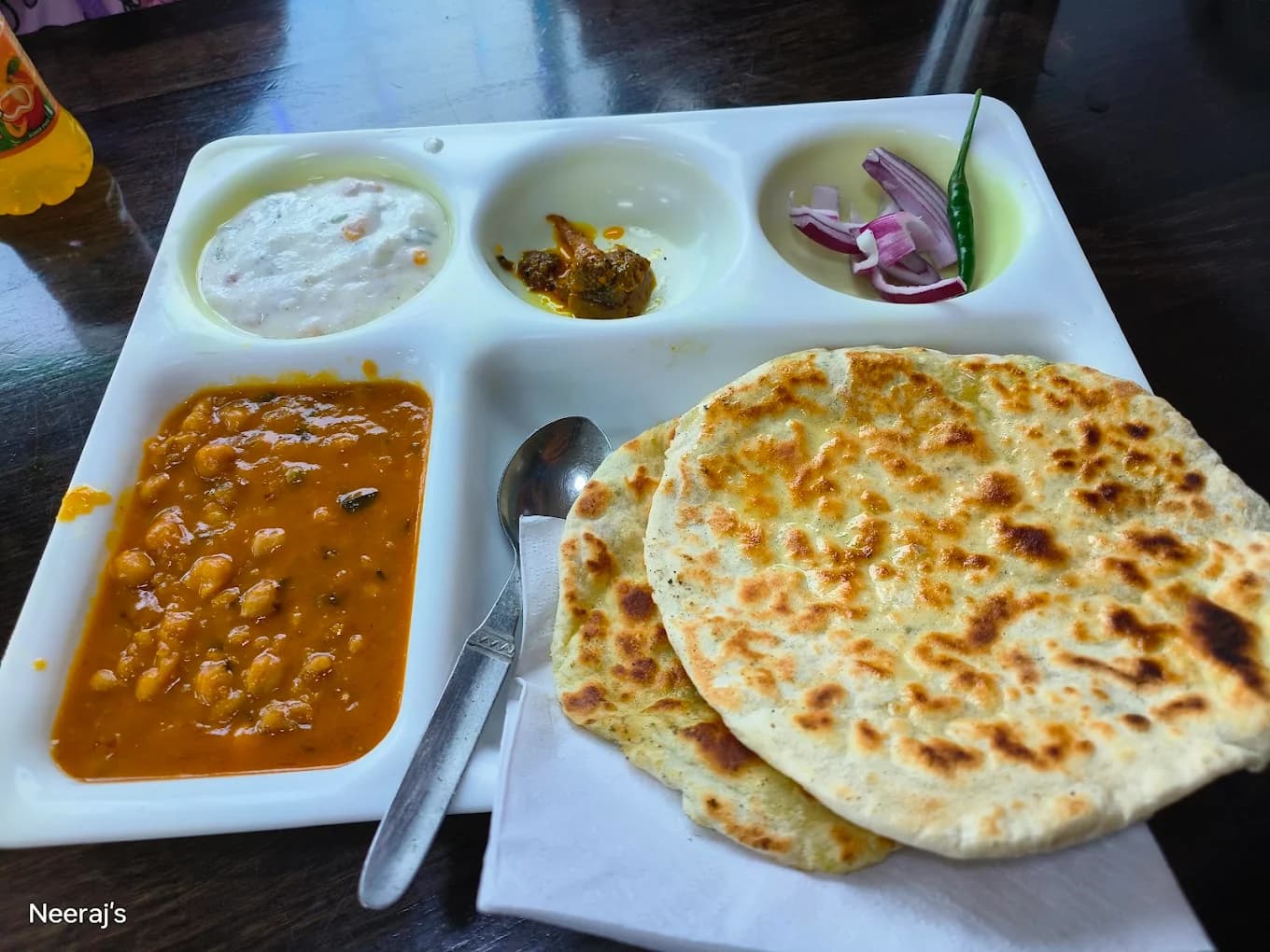 White partitioned tray containing chickpea curry, flatbread, raita, pickle, and sliced red onions.